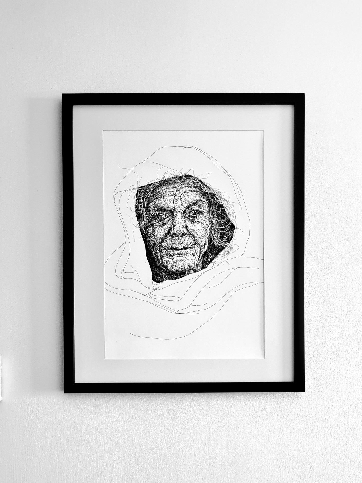 Framed black and white portrait of an elderly woman on a white wall. Her wrinkles are detailed scribbled pen lines surrounded by minimalist lines for clothing